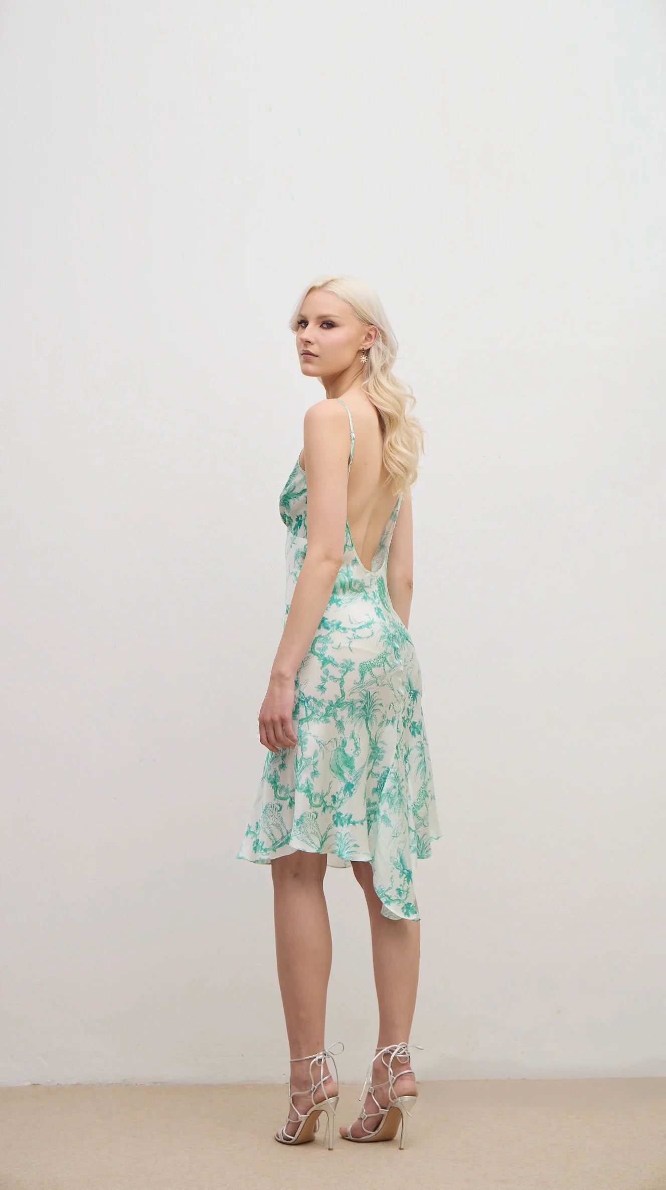 A blonde model wearing a green and white floral print midi dress with thin straps and a ruffled hem, paired with strappy high-heeled sandals, standing against a plain off-white studio wall.