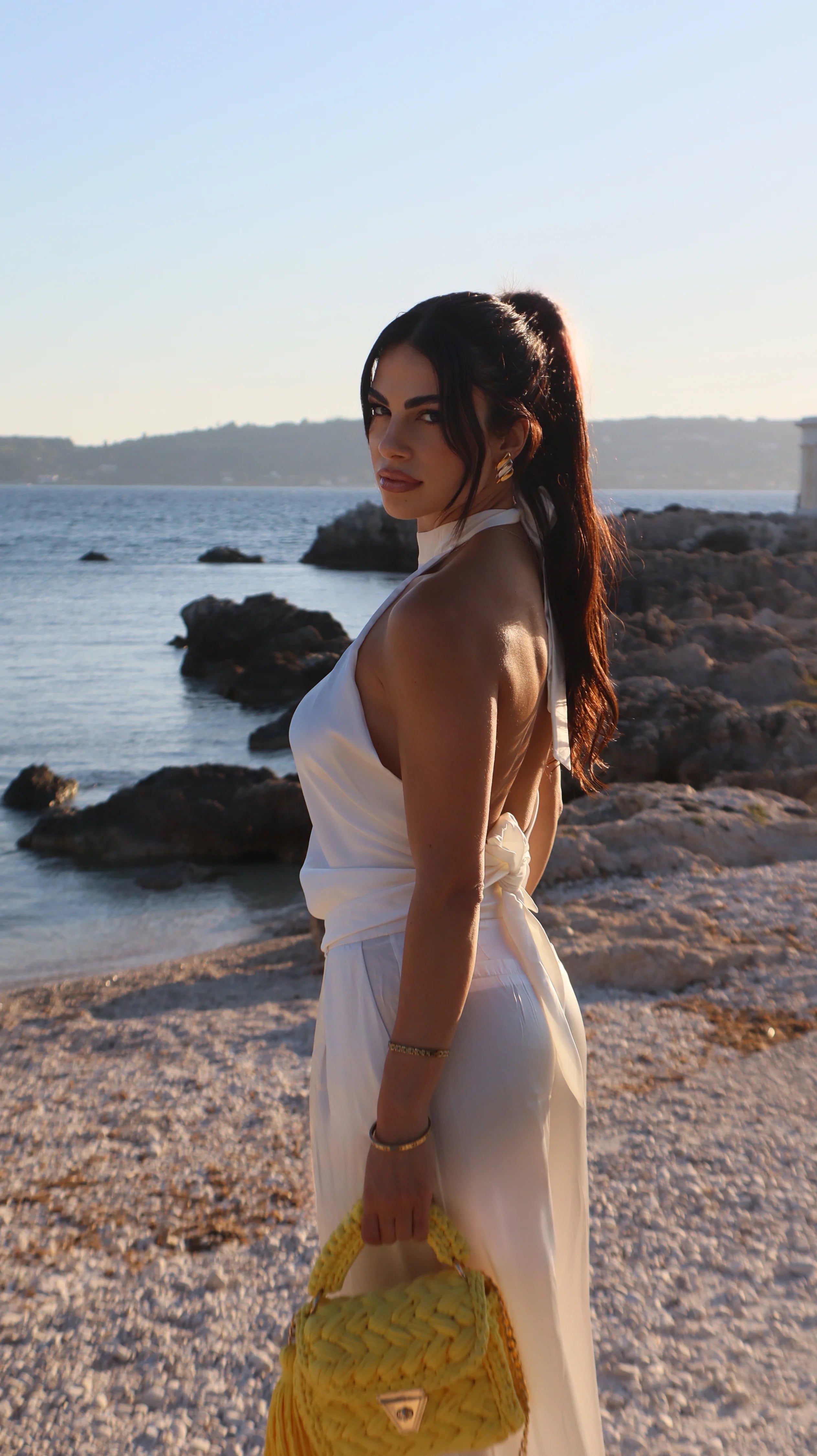 A woman stands on a rocky beach wearing a white halter-neck dress and holding a bright yellow woven handbag.