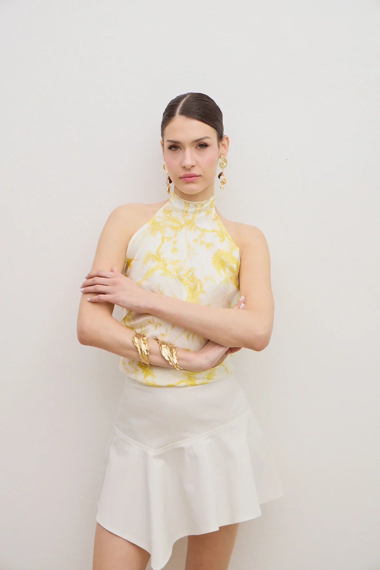 A woman modeling a sleeveless high-neck halter top with a yellow and white marble pattern, paired with a white ruffled mini skirt, against a plain light gray background.
